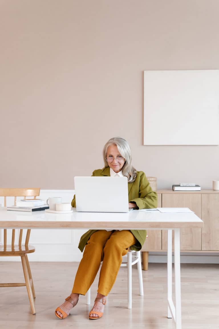 Full body aged woman in casual clothes sitting at table and using laptop while working on startup project in modern workplace