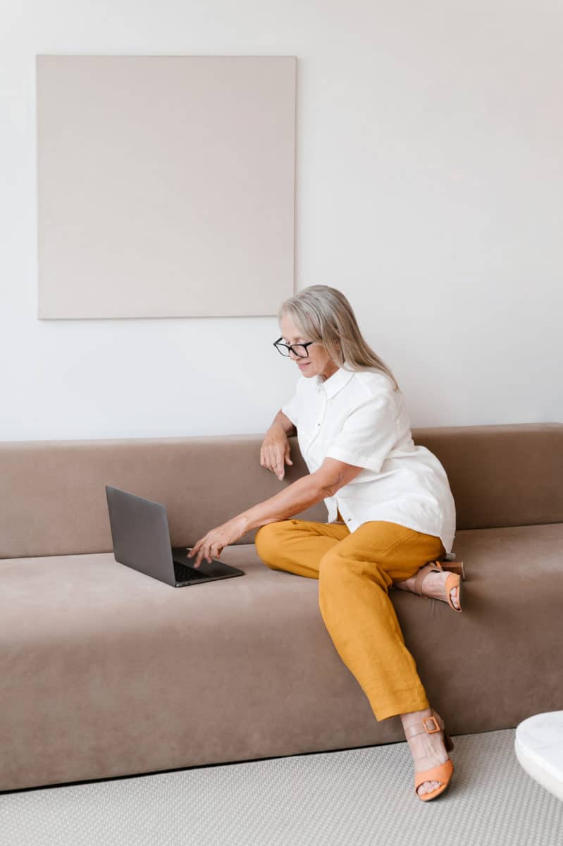 Full body side view of person in stylish clothes and eyeglasses sitting on sofa and browsing laptop in modern living room