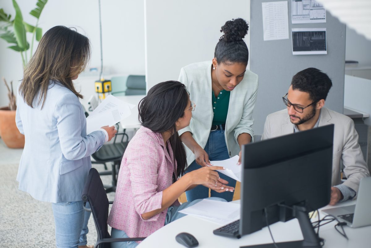 Four colleagues huddled around one side of the desk in front of a computer over a small stack of papers. | What is a Business Associate Agreement?