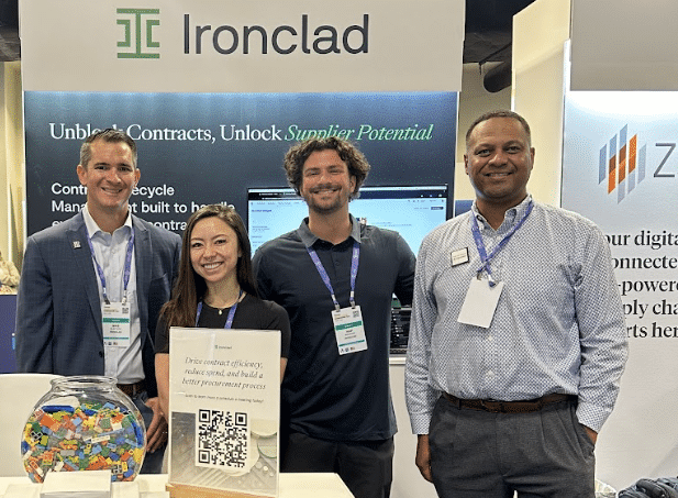 Four people stand smiling at an Ironclad booth during an event. A sign with a QR code and a bowl of colorful candy are on the table. Company banners are visible in the background.