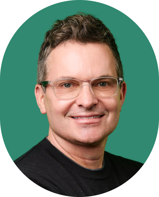 Smiling man with short brown hair, wearing clear glasses and a black shirt, posed in front of a solid green background.