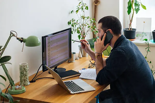 Person talking on a cellphone. He is sitting on a wooden desk at his home office and looking at computer screeens in front of him.