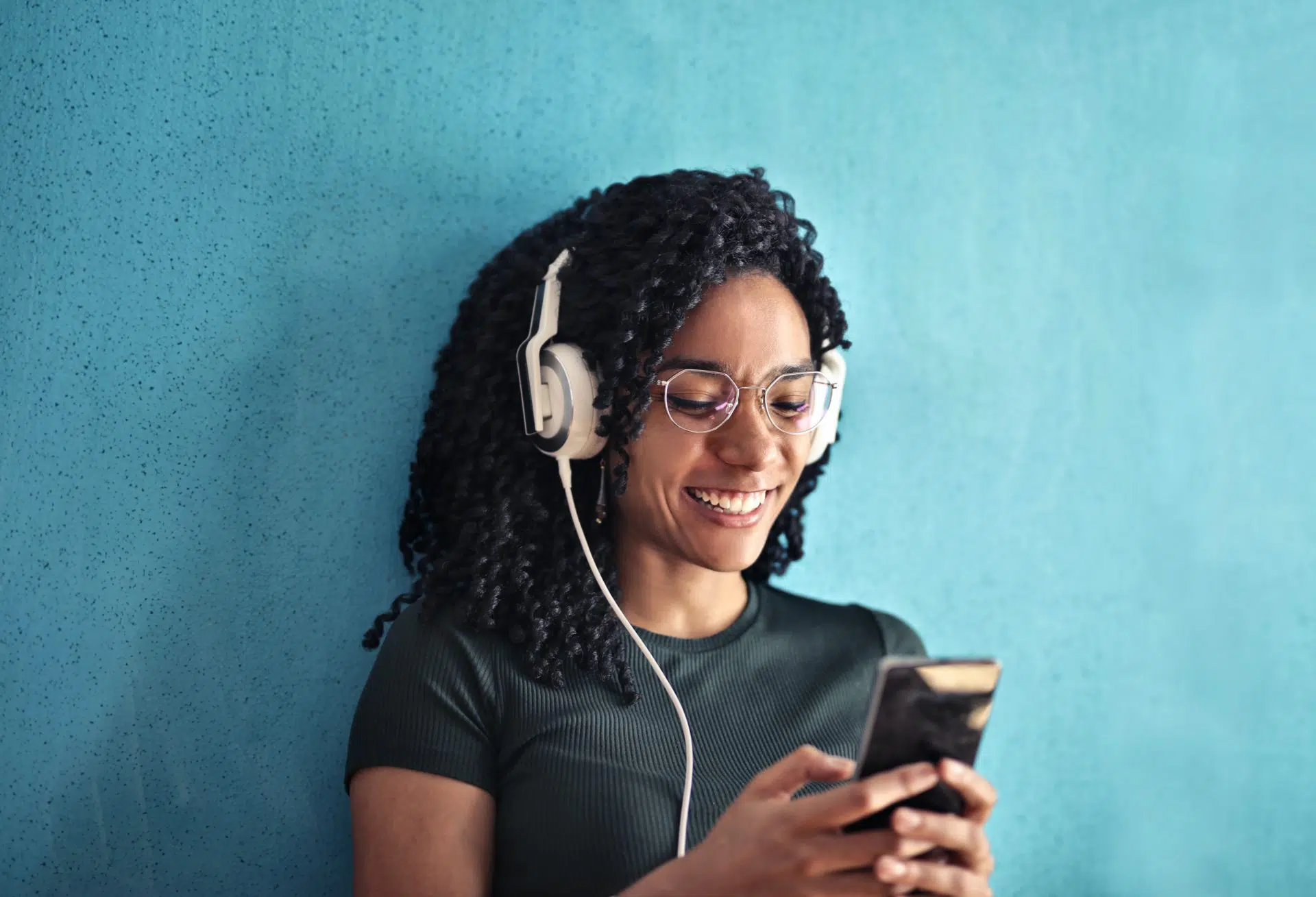 A smiling woman with curly hair and glasses wears large white headphones and looks at her smartphone while standing against a bright blue wall.