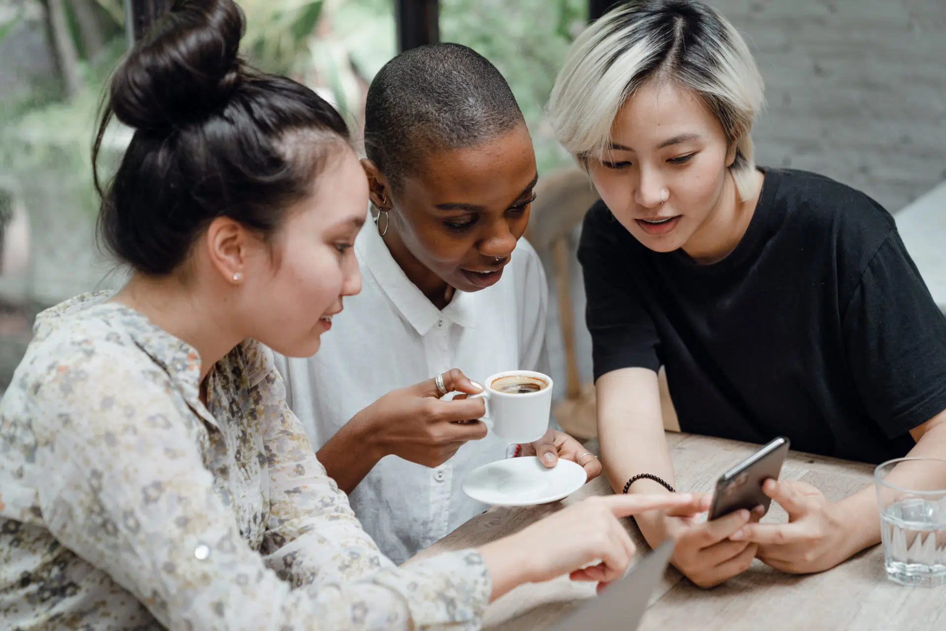 Three women sitting at a table, looking at a smartphone together. One woman holds a cup of coffee, while another points at the phone. They appear engaged and are in a bright, casual setting.
