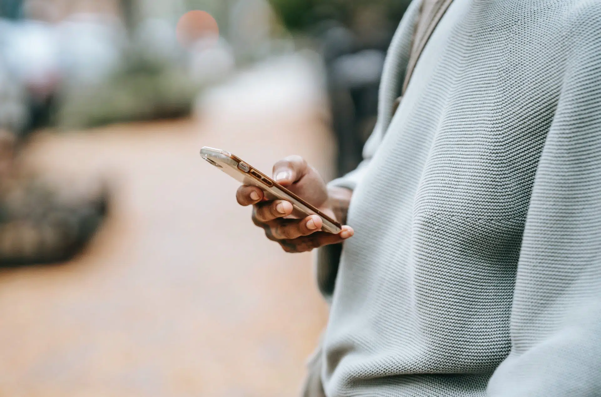 A person wearing a light gray textured sweater is holding and using a smartphone outdoors, with a blurred street scene in the background.