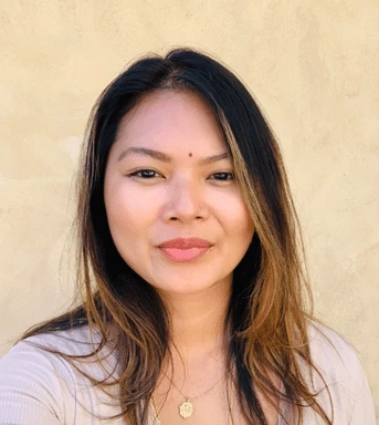 A woman with long, straight, dark brown hair and light highlights, wearing a beige top and a gold necklace, stands in front of a plain beige background. Her outlaw alternative style shines as she smiles softly at the camera.