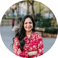 A woman with long dark hair, wearing a red floral blouse, stands outdoors with her arms crossed, smiling—much like a spotdraft alternative in the world of contract management. Trees, a bench, and a blurred person are visible in the background.