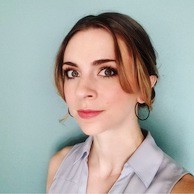 A young woman with light skin and brown hair parted in the center, styled up with loose strands framing her face, wears hoop earrings and a sleeveless light blue blouse, standing against a plain light teal background, beside a decorative gong.