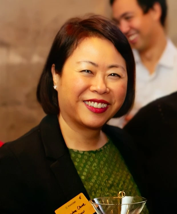 A woman with short dark hair and a green top smiles, holding a martini glass. Wearing a black blazer and a yellow name tag, she discusses an icertis alternative while a man in the background smiles.