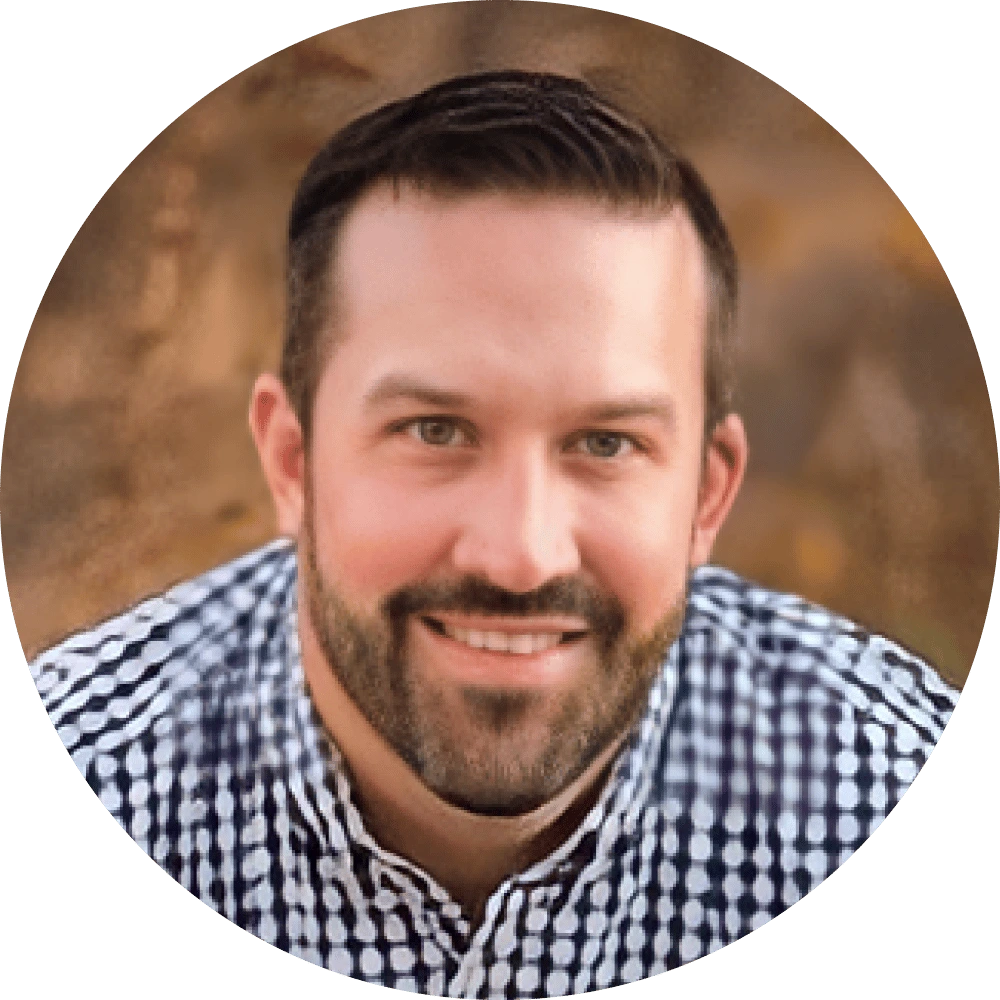 A man with short dark hair and a trimmed beard is smiling. He is wearing a black and white checkered shirt, posed in front of a blurred brown-toned background, while considering an Icertis alternative for contract management.