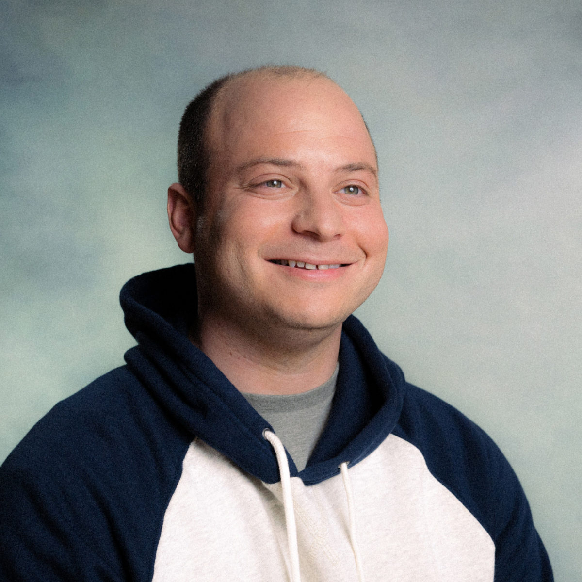 A smiling man with a receding hairline wears a navy and white demostack hoodie against a softly blurred, light-colored background.