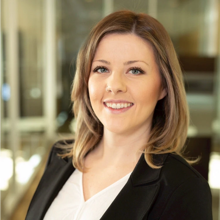 A woman with shoulder-length light brown hair, wearing a black blazer over a white top, smiles while standing in a modern office with glass walls, representing a team introducing a ContractPodAI alternative.