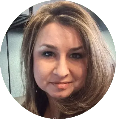 A woman with long, light brown hair and hazel eyes is looking at the camera with a slight smile. She is indoors, possibly exploring an agiloft alternative in a modern office setting.