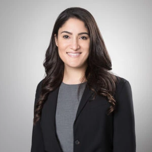 A woman with long dark hair, wearing a gray top and a black blazer, smiles at the camera against a plain light gray background, showcasing the professional vibe of a spotdraft alternative.