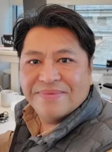 A man with short dark hair and a slight smile is pictured indoors, wearing a gray jacket over a collared shirt. The background, suggesting a professional space, hints he might be exploring a ContractWorks alternative for improved workflow.