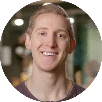 A young man with short, light brown hair smiles at the camera. He is wearing a casual shirt in an indoor setting with blurred lights and people in the background, perhaps discussing the best SpotDraft alternative for contract management.