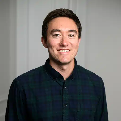 A smiling young man with short dark hair wearing a dark green plaid button-up shirt stands in front of a plain light-colored wall.