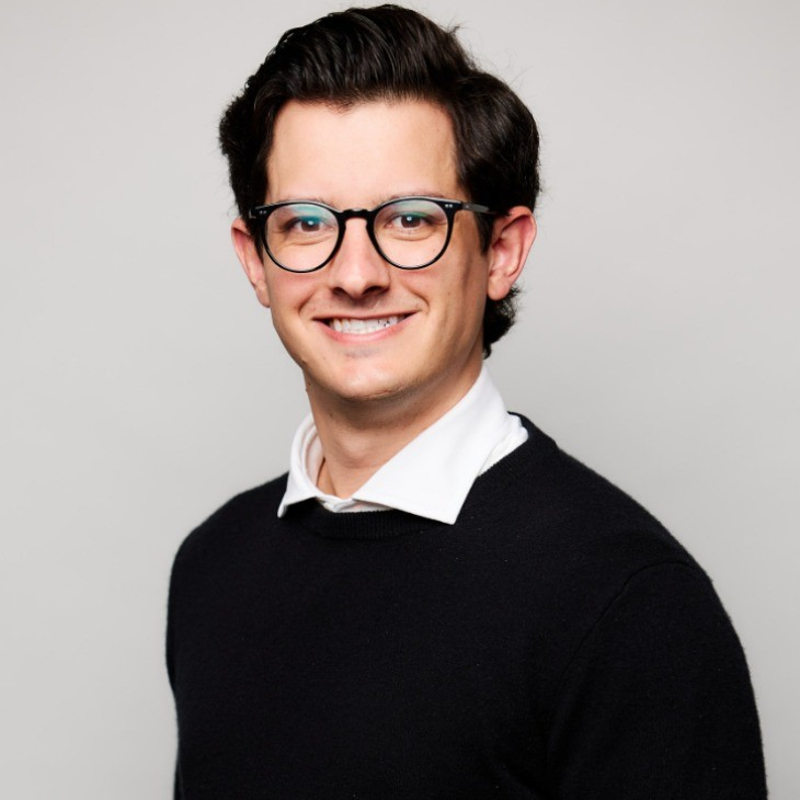 A young man with short dark hair and glasses smiles at the camera. He is wearing a white collared shirt under a black sweater, standing against a plain light gray background.