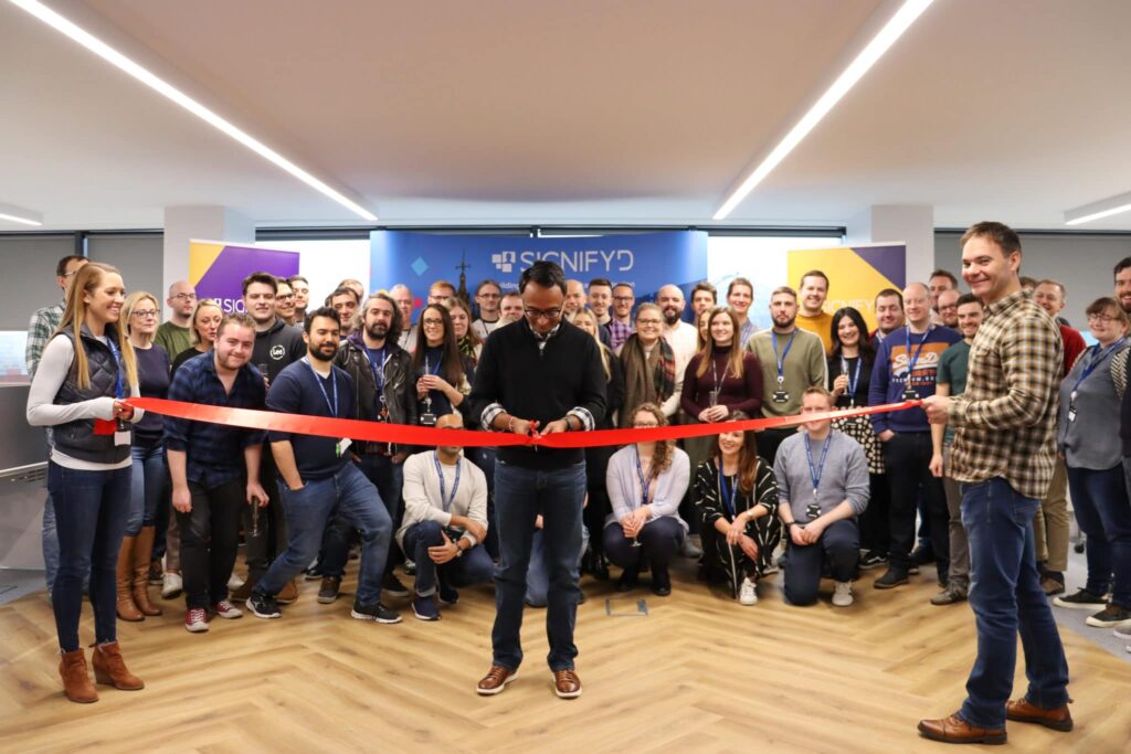 A group of people gathers and smiles as a man in the center cuts a red ribbon, held by two others, during an indoor ribbon-cutting ceremony. A blue sign reading SIGNIFYD is visible in the background.
