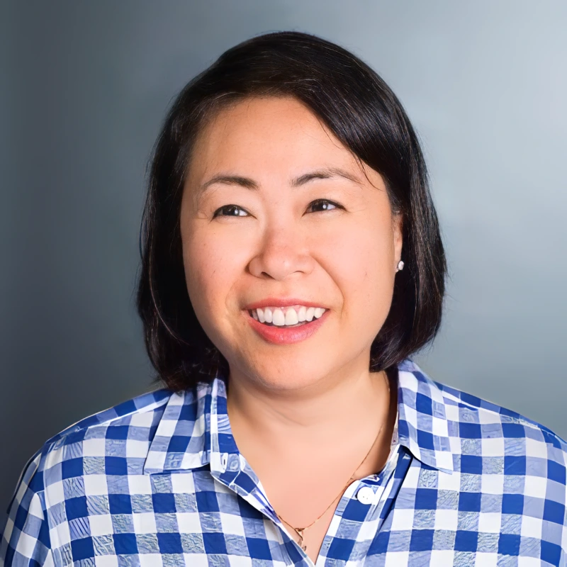 A woman with short black hair smiles while wearing a blue and white checkered shirt, in front of a plain gray background—capturing the approachable vibe you'd expect from a leading SpotDraft alternative.