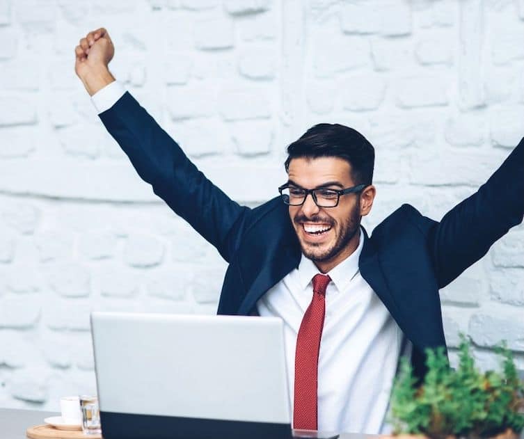 A man in a suit and red tie sits at a desk with a laptop, smiling broadly and raising both arms in excitement or celebration.