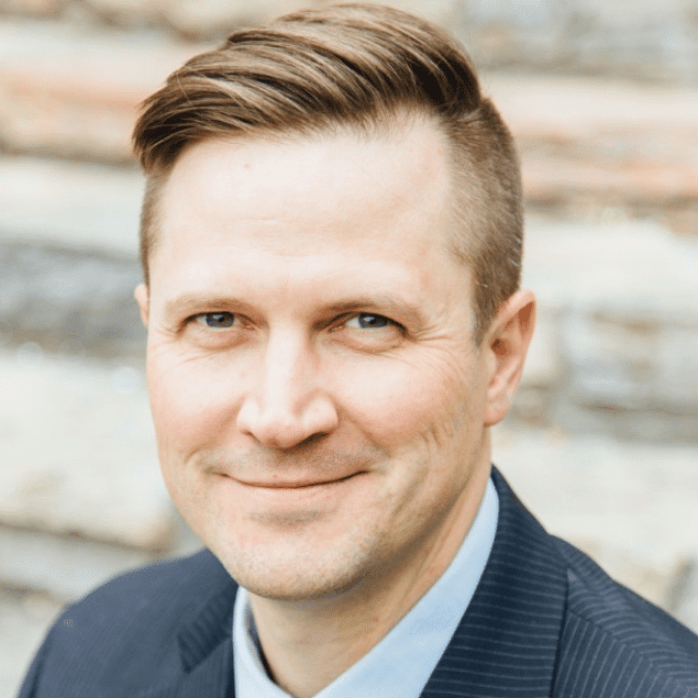 A man with short, light brown hair styled to the side, wearing a pinstripe suit and collared shirt, smiles at the camera—ideal for a professional seeking a linksquares alternative. The background features blurred stonework.