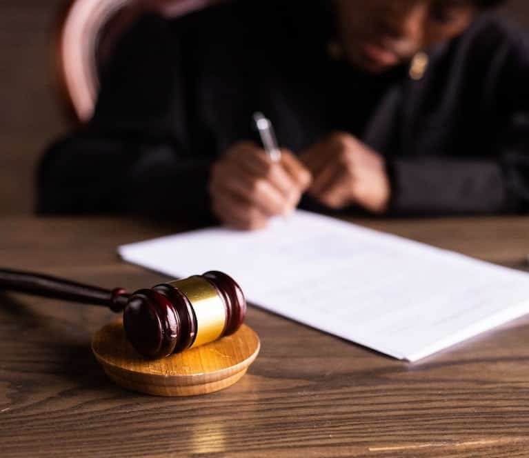 A wooden gavel rests on a block in the foreground, while a person in dark clothing writes on a document at a desk in the blurred background.