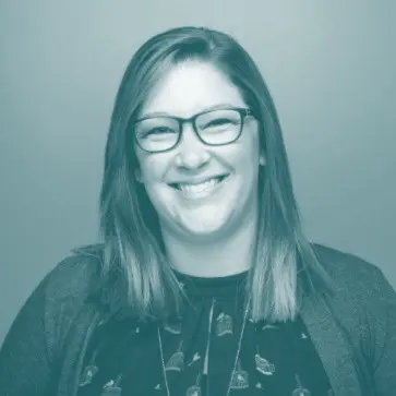 A woman with straight, shoulder-length hair and glasses smiles at the camera. She is wearing a patterned top and a cardigan, with a plain background behind her. The image has a blue-tinted filter.