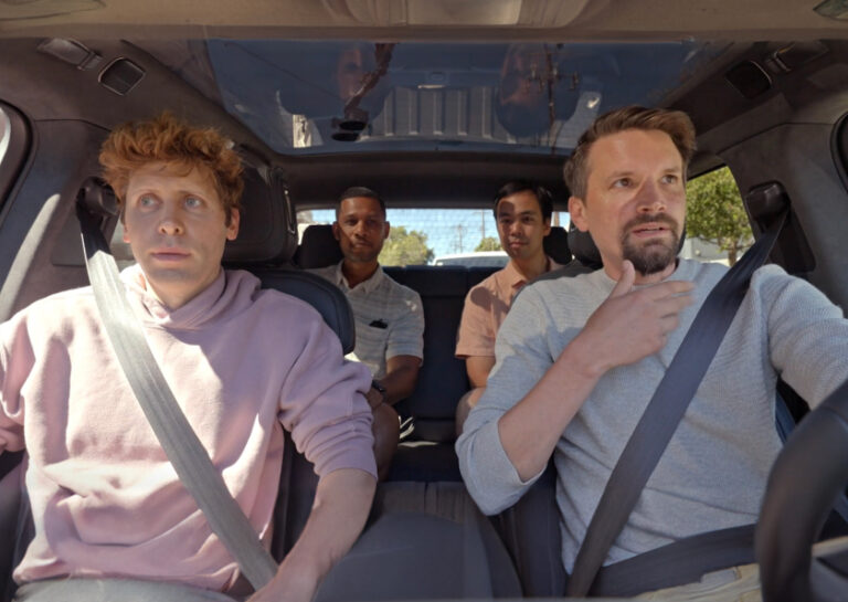 Four men sit inside a car; two in the front seats and two in the back. The driver has one hand on the wheel and the other on his chin, while the others look forward with neutral expressions.