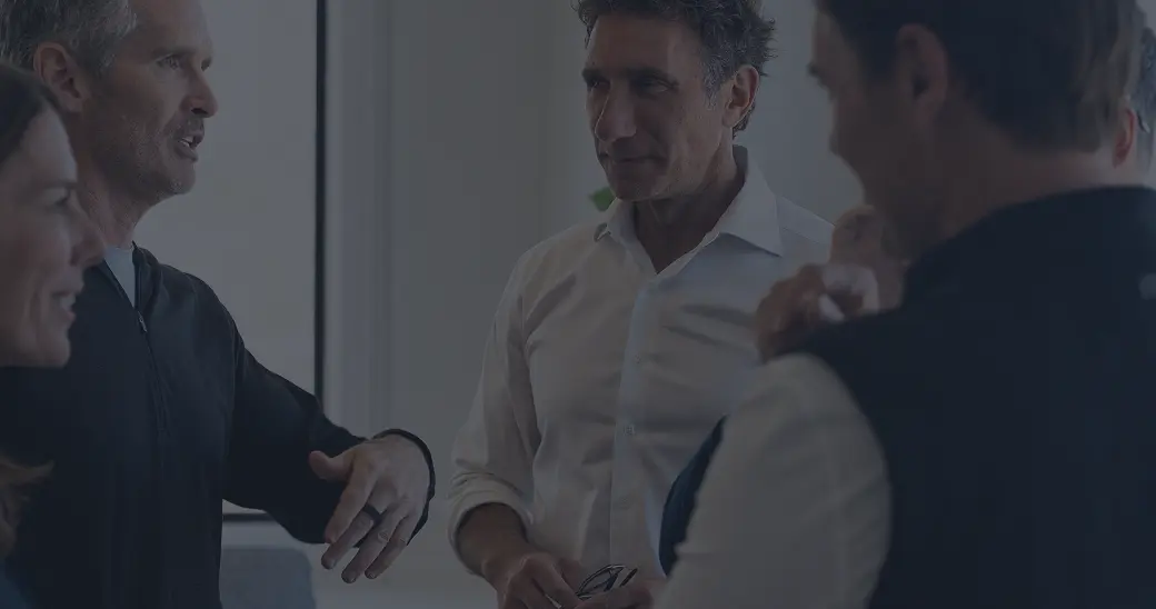 Four people are standing indoors, engaged in conversation. One man gestures with his hands while others listen attentively, creating a collaborative and professional atmosphere.