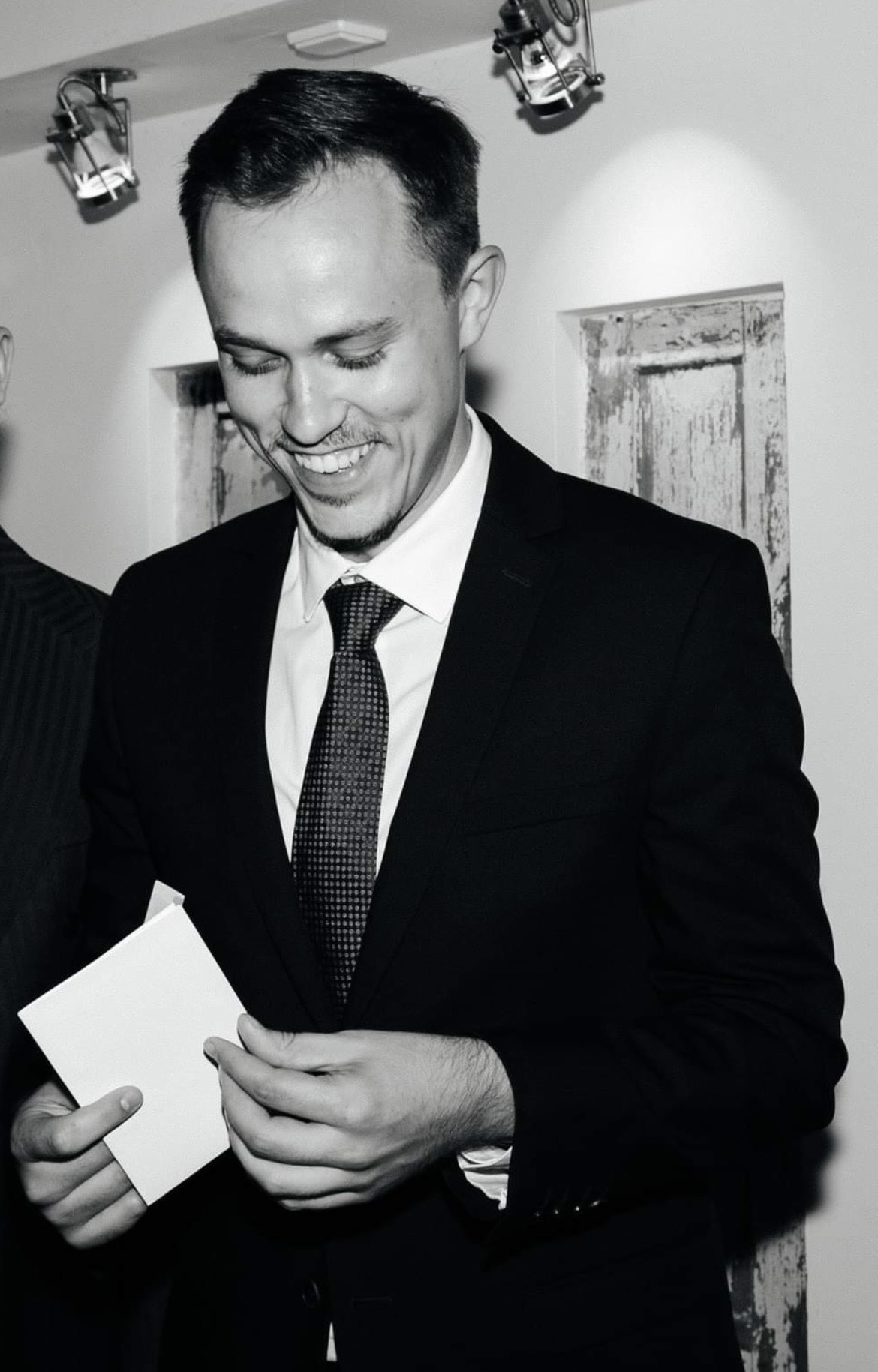 A man in a suit and tie smiles while holding an envelope, standing indoors against rustic wooden panels. The black and white photo evokes a timeless charm, reminiscent of vintage snapshots found on Vinted.