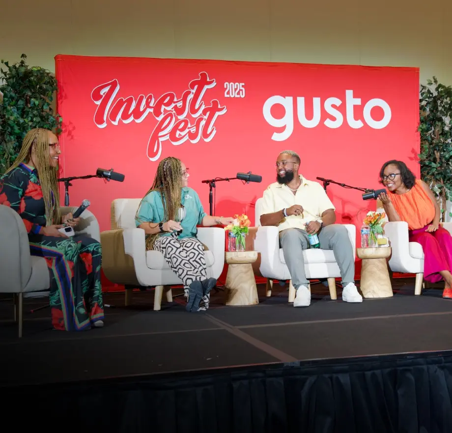 Four people sit on stage in front of a red Invest Fest 2025 gusto backdrop, having a panel discussion with microphones, surrounded by plants and decorative tables with flowers.