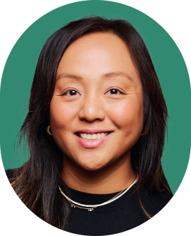 A woman with long dark hair smiling, wearing a black top, gold hoop earrings, and layered necklaces, shown against a green background in an oval frame.