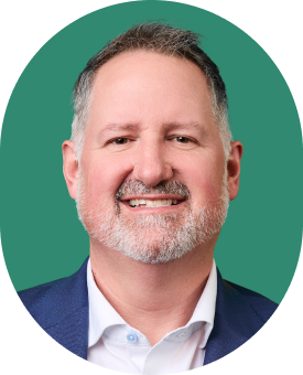 A middle-aged man with short gray hair and a beard, wearing a blue suit jacket and white shirt, smiling in front of a green background.
