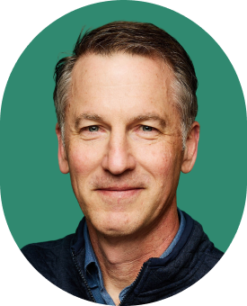 A middle-aged man with short gray hair and blue eyes, wearing a navy jacket over a blue shirt, smiles softly against a solid green background. The photo is cropped in an oval shape.