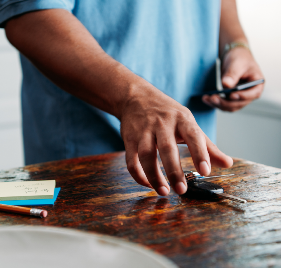 A person in a blue shirt reaches for car keys on a wooden table, with a phone, pencil, and sticky notes also visible.