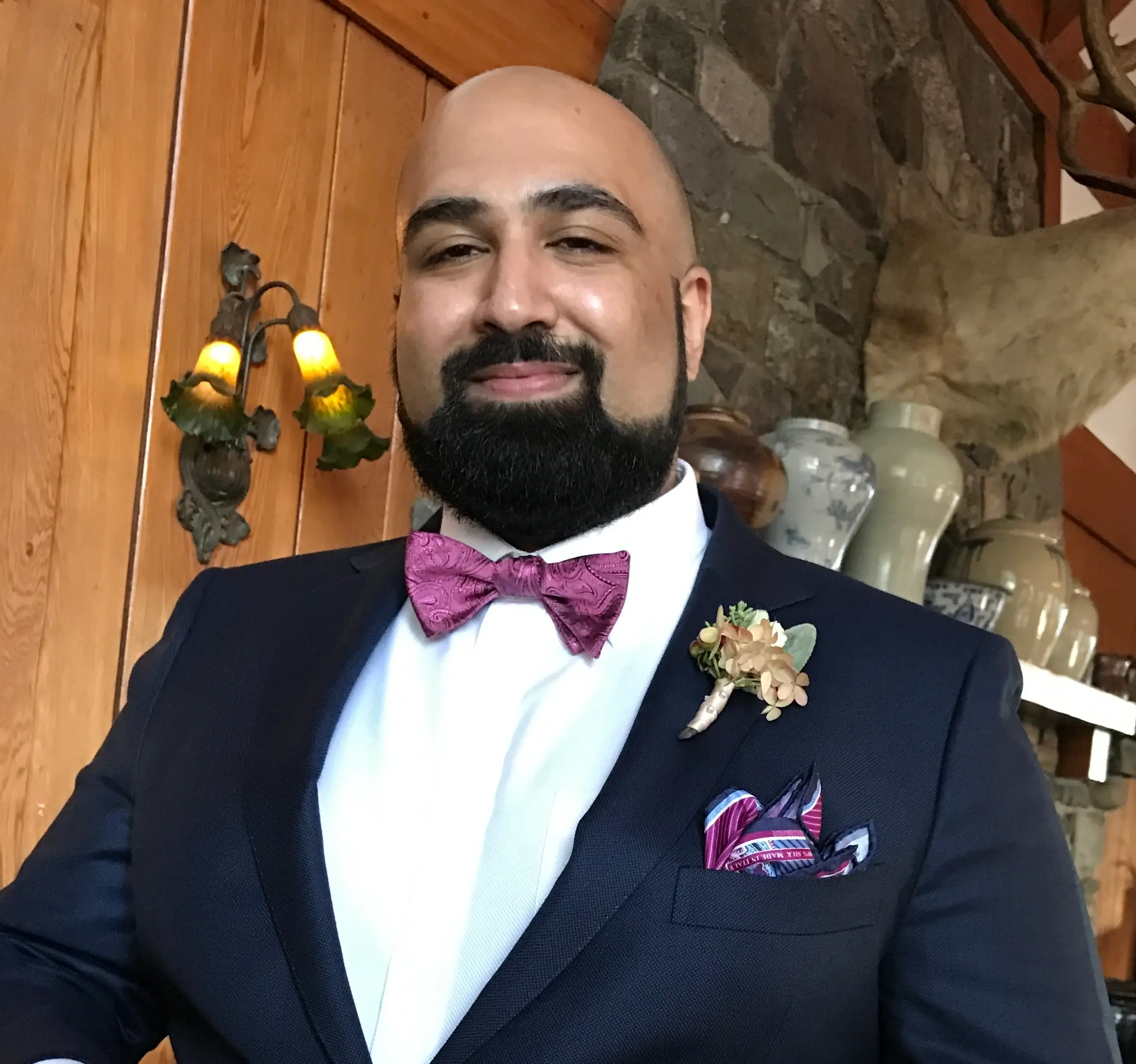 A man with a beard and bald head wears a navy suit, purple bow tie, boutonnière, and pocket square, standing indoors beside wooden walls painted in a warm Benjamin Moore shade and decorative vases.