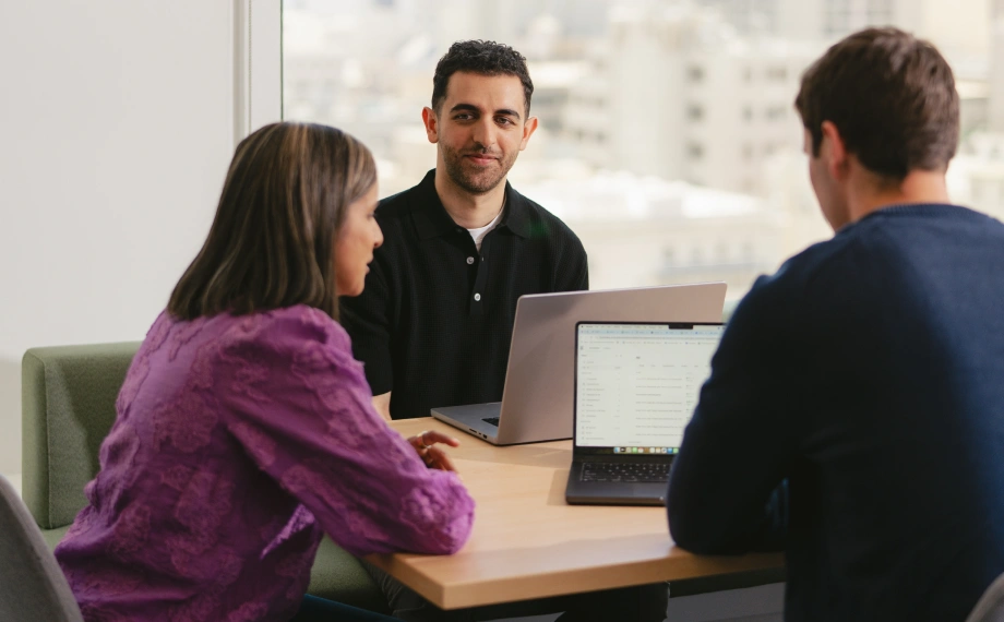 Three people sit at a table with laptops in a bright office, engaged in discussion about AI legal research. Two face away from the camera, while one faces forward, smiling. City buildings are visible through the window behind them.