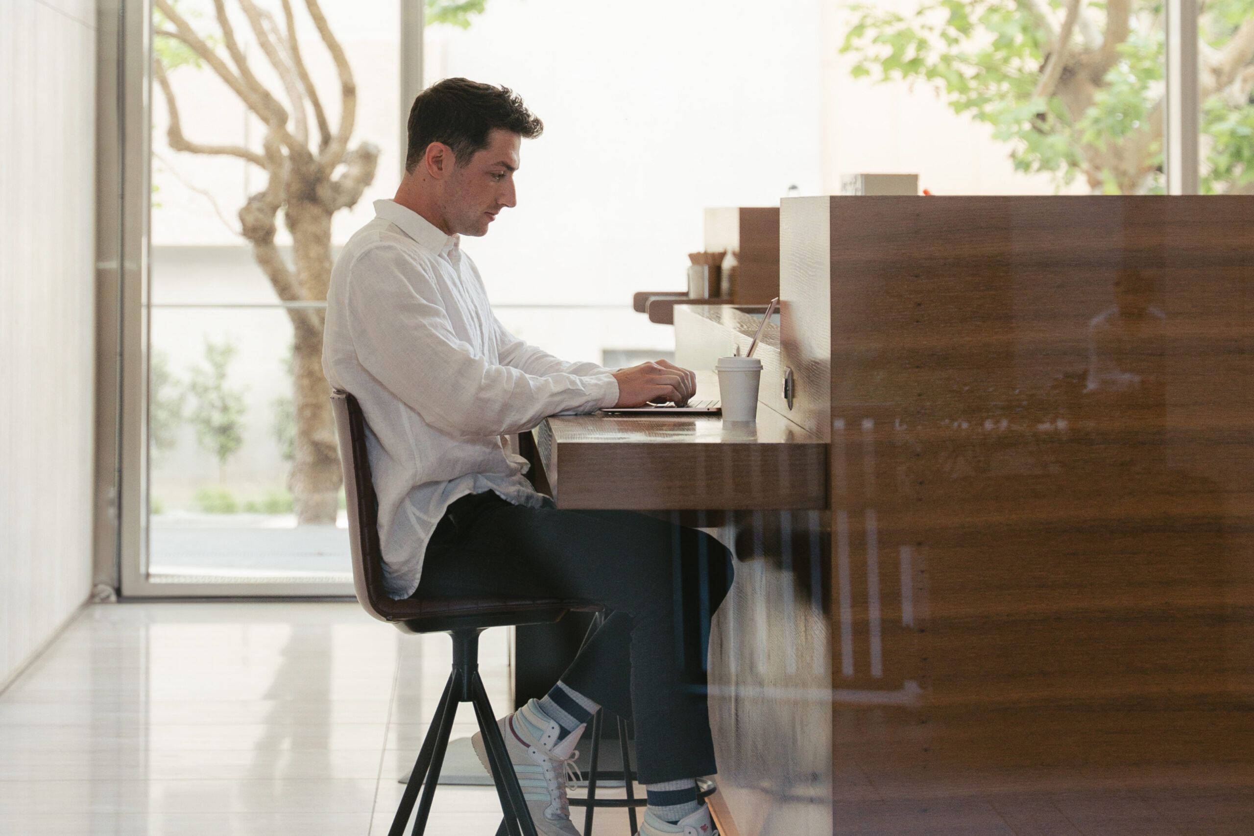 A man in a white shirt sits on a bar stool at a wooden counter, working on a laptop with a drink next to him. The setting is bright, with large windows and trees visible outside.