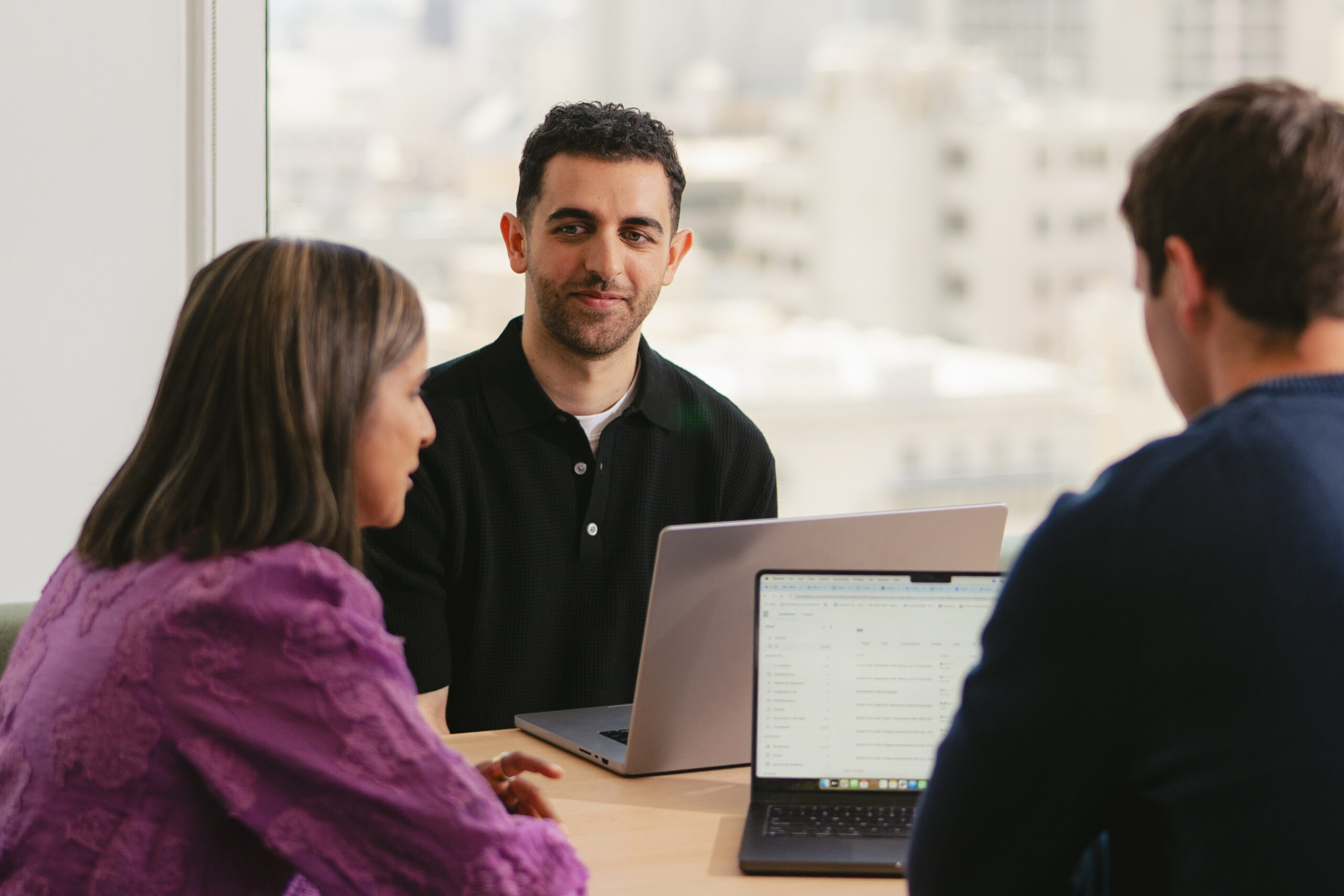 Three people sit around a table with laptops in an office, engaged in conversation. One person faces the camera while the other two have their backs turned. A cityscape is visible through the window behind them.