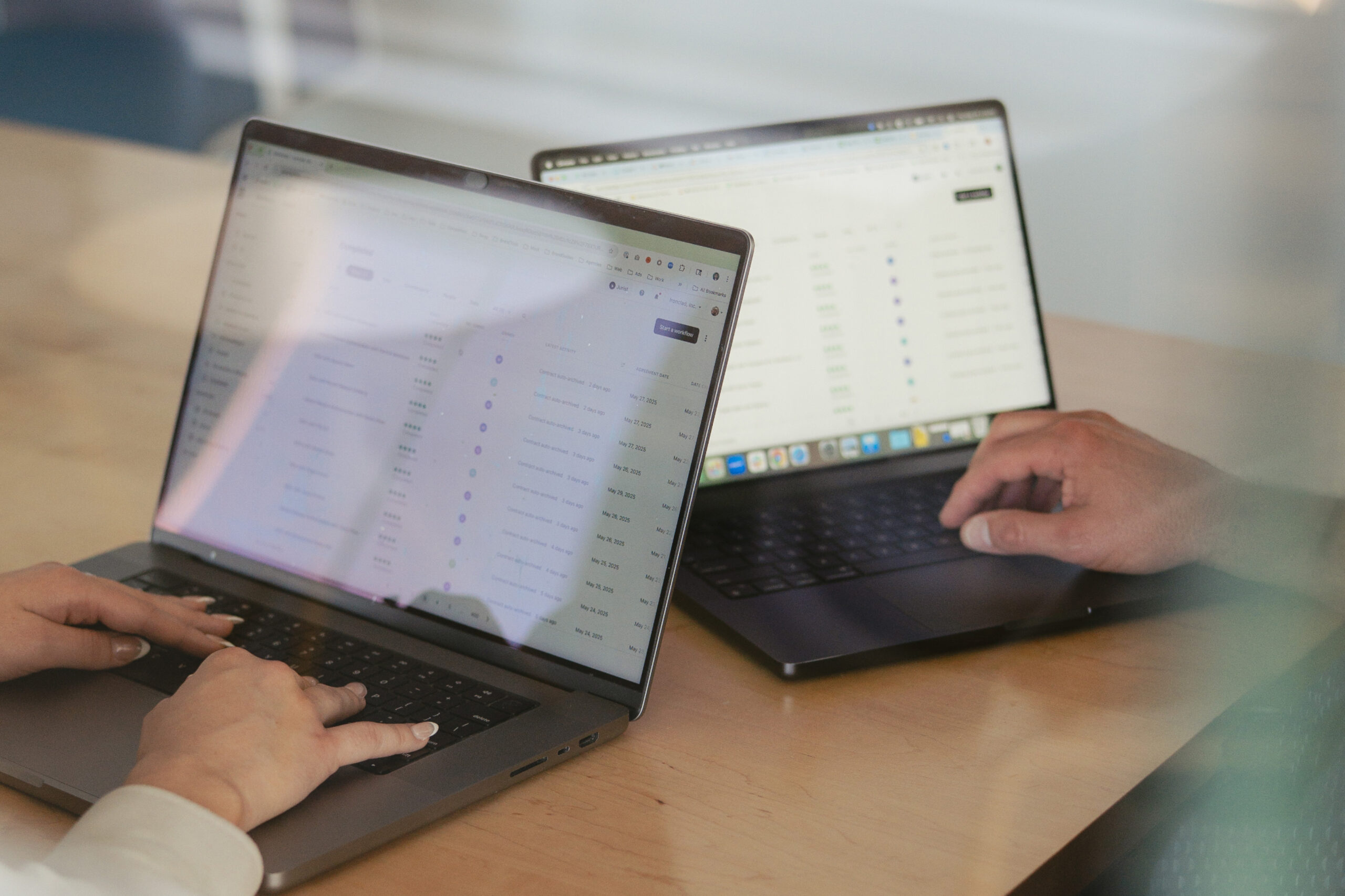 Two people sit at a table working on laptops, with data and charts displayed on their screens. The image is taken through a reflective surface, slightly blurring some details.