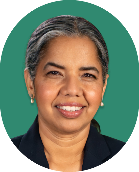 A woman with gray-streaked dark hair pulled back, wearing a dark blazer and pearl earrings, smiles in front of a solid green background.