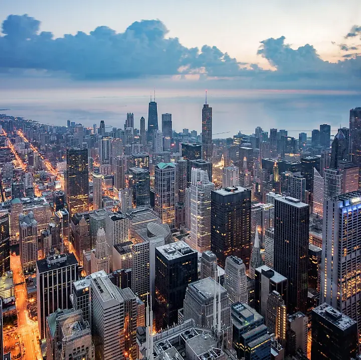 Aerial view of a city skyline at dusk with tall skyscrapers, illuminated streets, and a large body of water in the background under a cloudy sky.