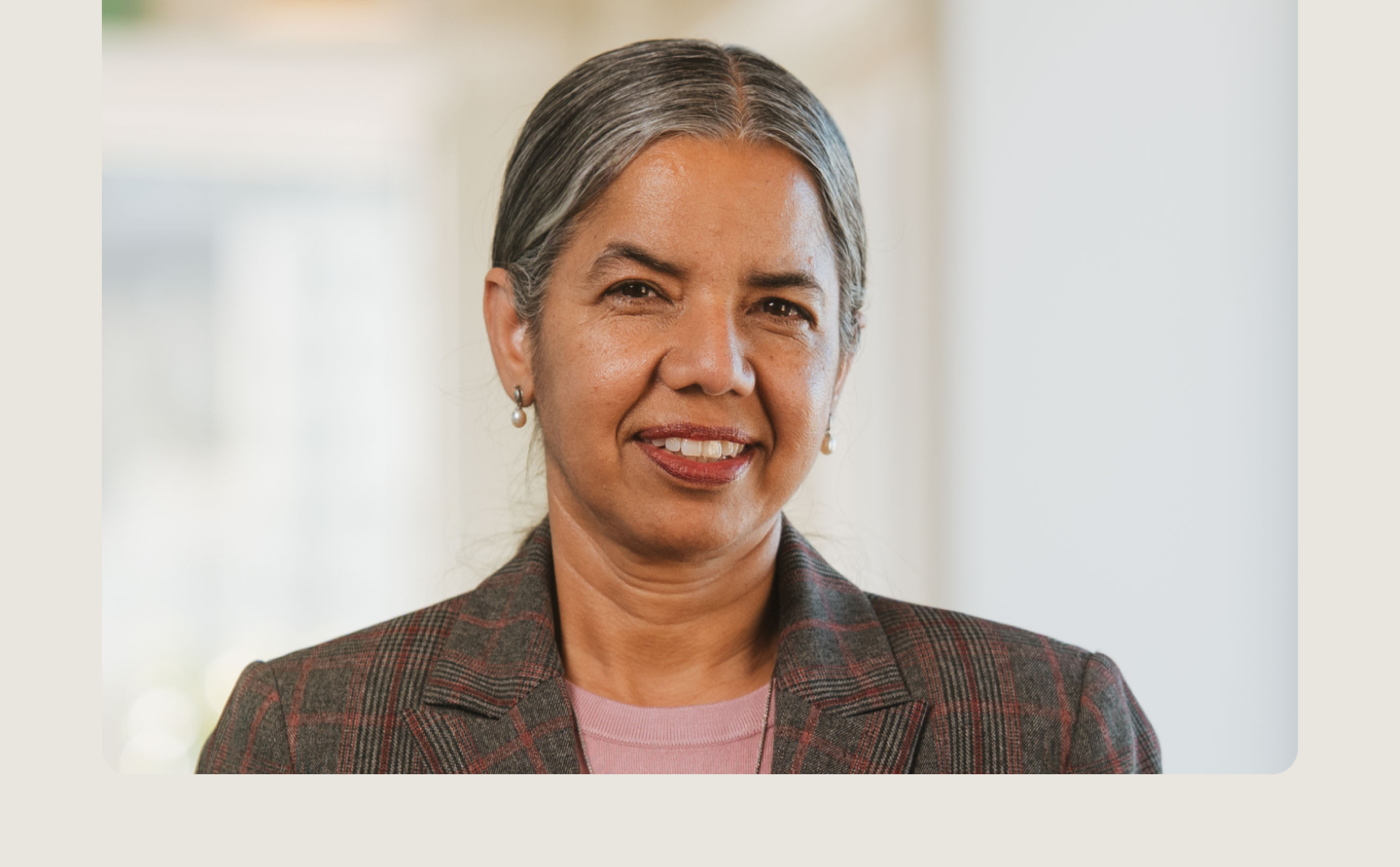 A woman with gray hair pulled back, dressed as a distinguished jurist in a brown plaid blazer and pink top, smiles at the camera against a softly blurred indoor background.