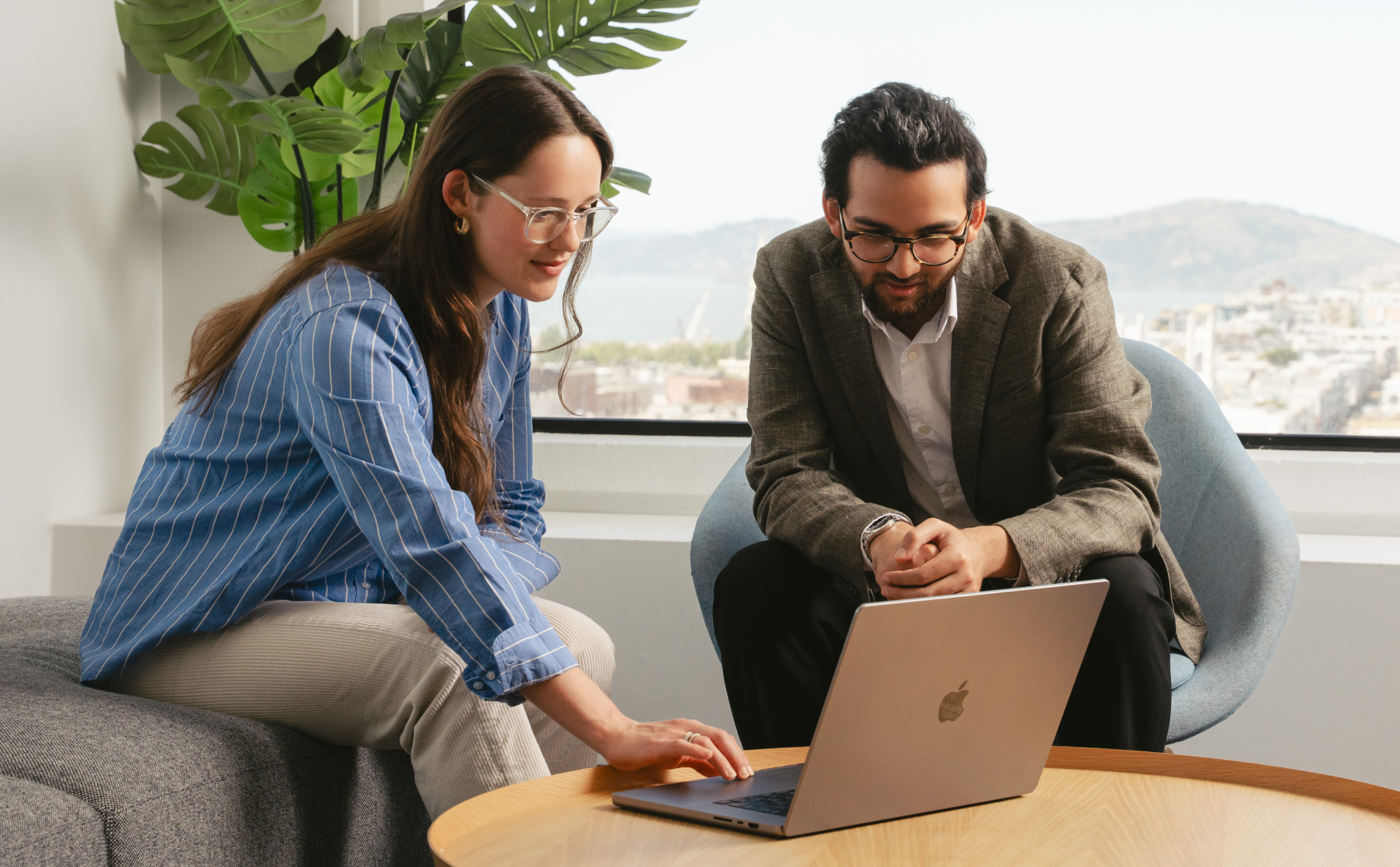 Two people sit in a modern office by a window, looking at a laptop on a wooden table. One points at the screen, sharing procurement horror stories, while the other listens intently. Green plants and city buildings frame the background.