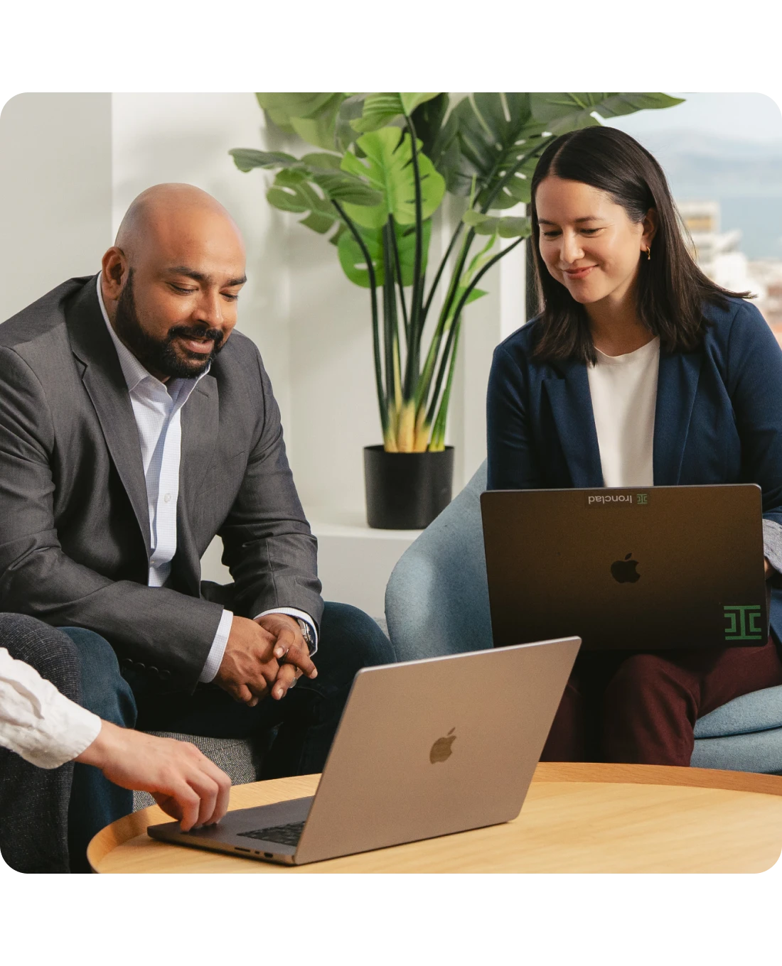 Two people sit together at a round table, both looking at open laptops. They appear to be having a discussion or meeting. There is a large plant in the background and natural light coming through a window.