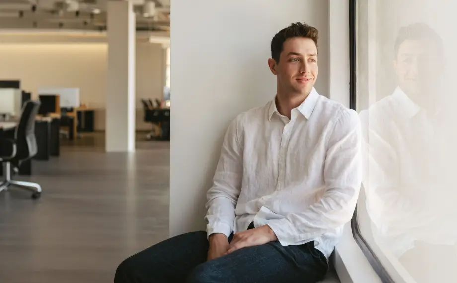 A contract manager in a white shirt sits by a large window in a modern office, looking outside with a slight smile. The office is spacious, with empty desks and chairs in the background.