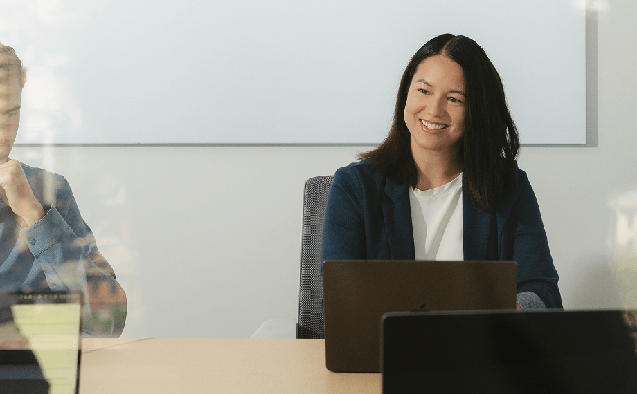 A woman with dark hair sits at a table in a meeting room, smiling and working on a laptop—possibly reviewing adhesion contracts examples—with another person partially visible on the left.