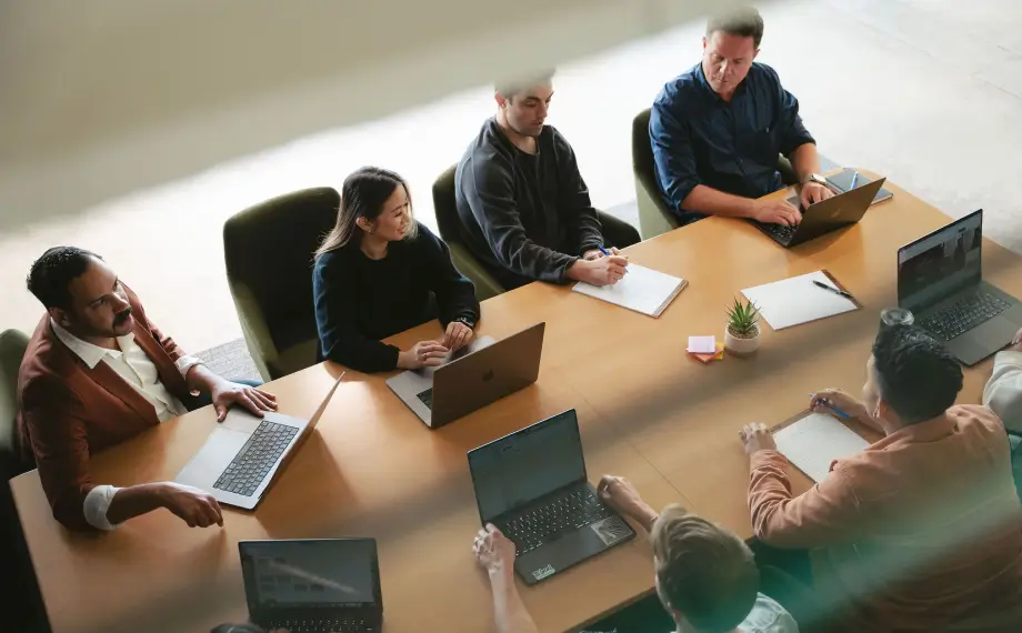 Seven people sit around a conference table with laptops, notebooks, and papers, engaged in a meeting or discussion focused on legal operations. The setting appears to be a modern office, viewed from above.