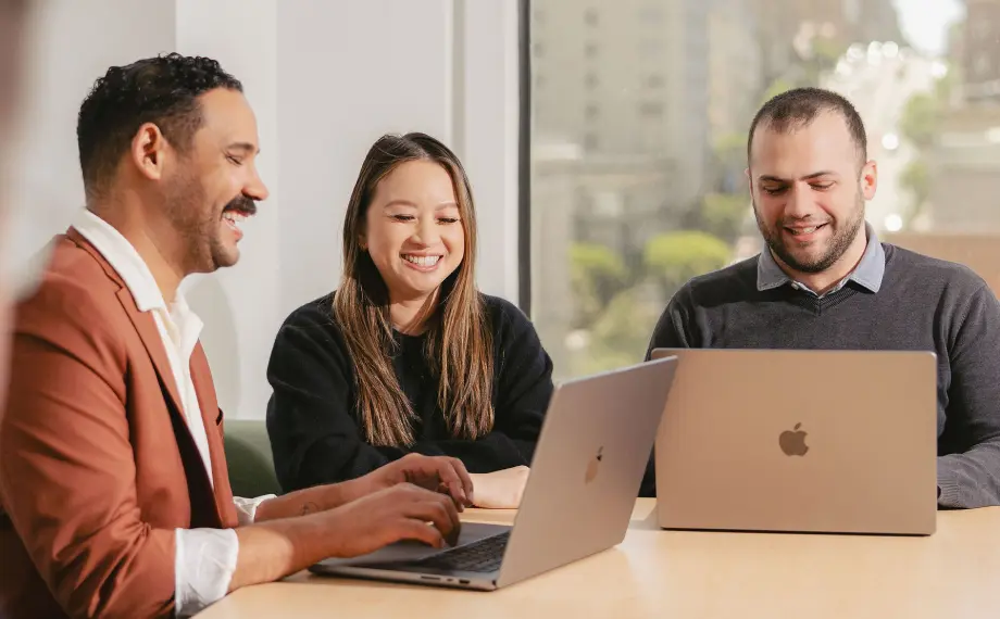 Three people sit at a table with laptops, smiling and working together in a bright office with large windows showing a cityscape outside. Their collaboration highlights how contract management in Salesforce is a win for both sales and legal teams, streamlining their workflow in an inspiring environment.